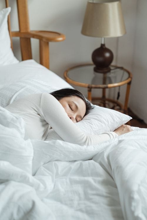 A young woman sleeping comfortably in a cozy bed with white bedding and natural light